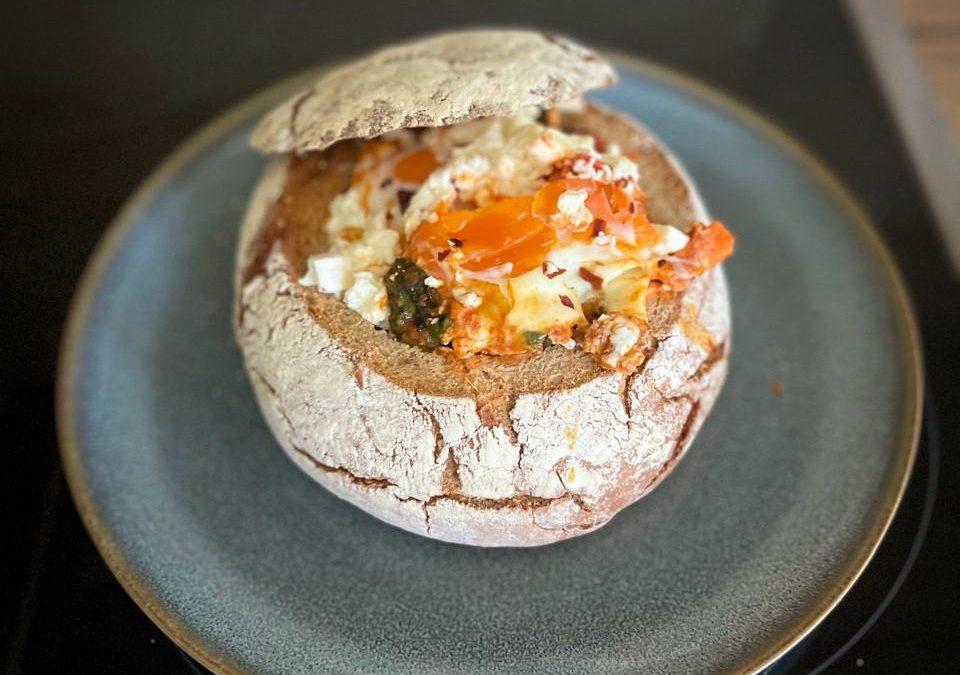 Imagem de uma shakshuka nutritiva e colorida com batata doce, espinafre, linguiça defumada e ovos, servida dentro de um pão redondo